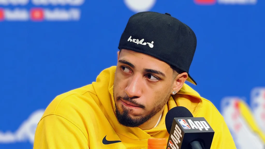 Tyrese Haliburton, wearing a black baseball cap with white writing and a yellow hoodie, looks to his right while speaking into a microphone. The background is a blurred blue.