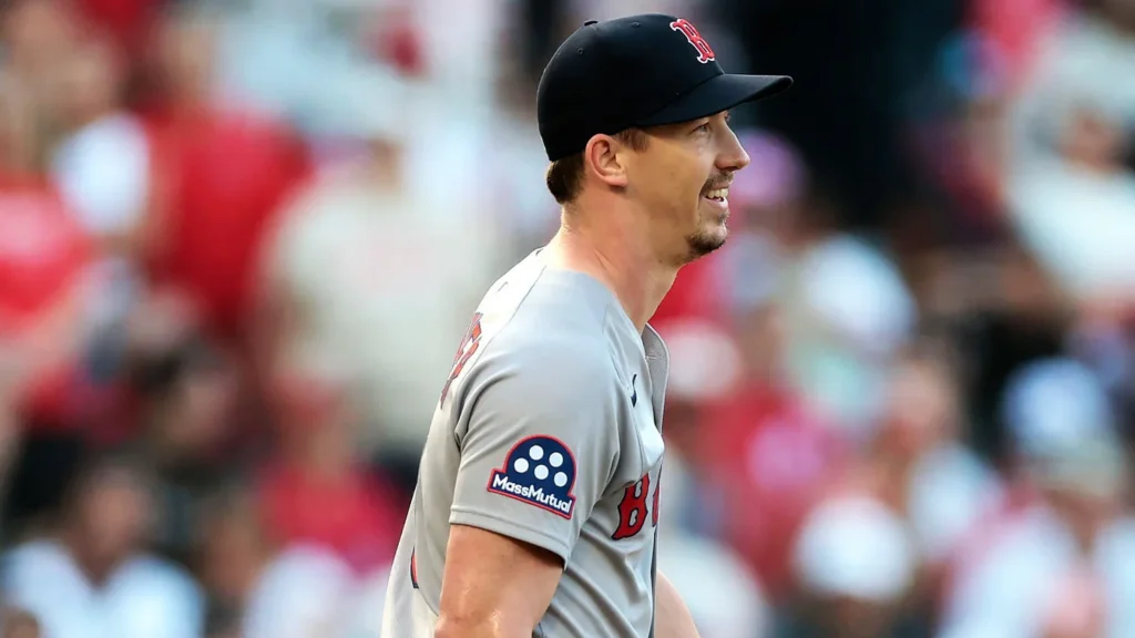 A close-up, side profile of Boston Red Sox pitcher Walker Buehler on the field, wearing a gray jersey with a red "B" logo and a black baseball cap. He is smiling and looking to his right. The background is blurred with hints of red and green.