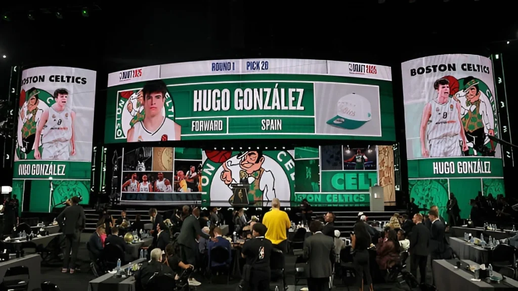A large screen at the NBA Draft shows a graphic for Hugo González, a forward from Spain, being drafted by the Boston Celtics as the 28th pick in the first round.