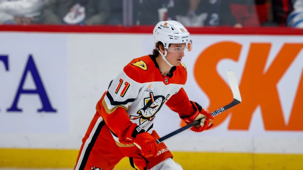 Trevor Zegras in his orange and white Anaheim Ducks jersey on the ice, holding a hockey stick.