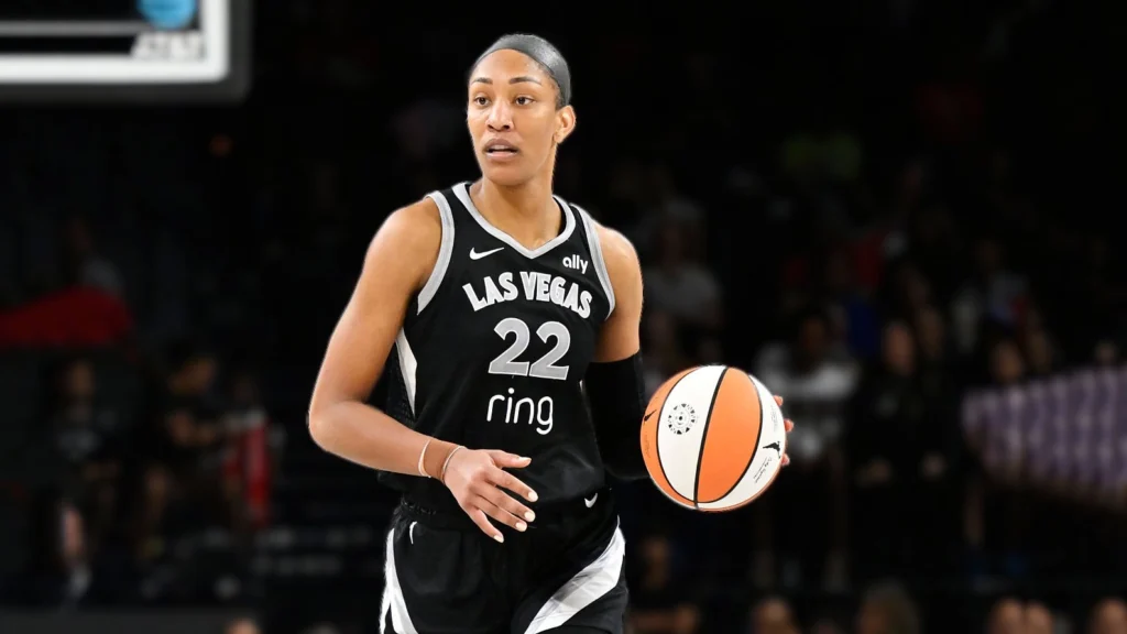 A'ja Wilson, an athletic female basketball player in a black Las Vegas Aces uniform (jersey number 22) and an arm sleeve, is focused on dribbling a basketball with her right hand. She is on a basketball court with a blurred, dark background suggesting a crowd.