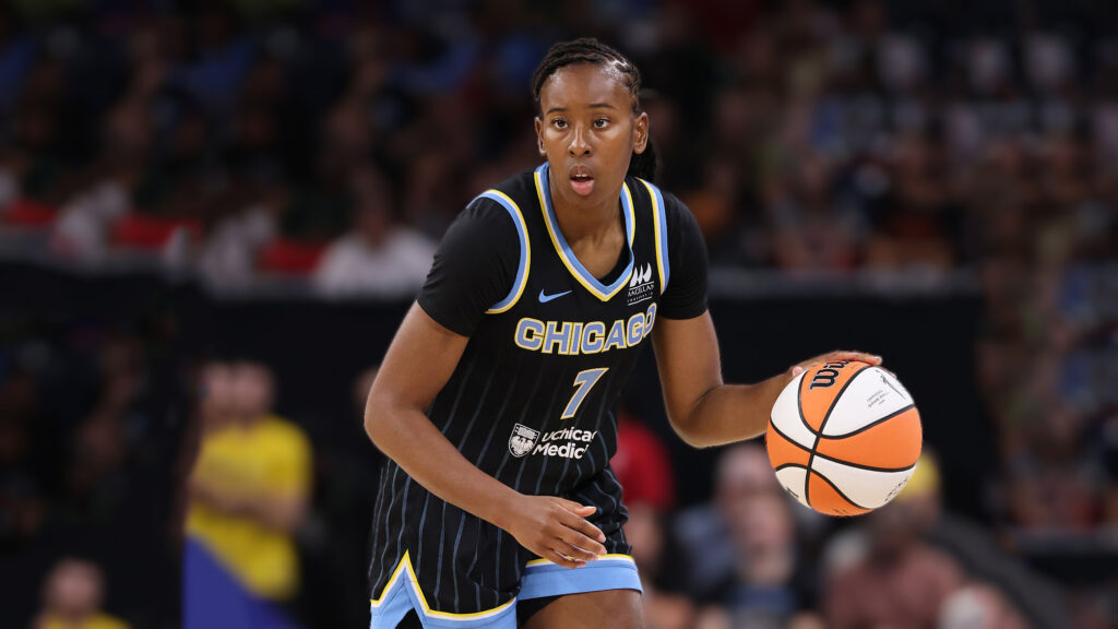 A female basketball player, Ariel Atkins, wearing a black Chicago Sky jersey with the number 7, dribbles an orange and white basketball. She has braids and is looking forward with a focused expression. The background is blurred, showing a sports arena.