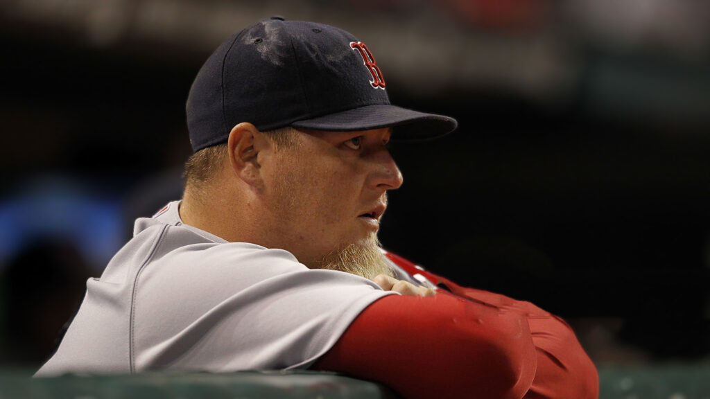 A side profile shot of Bobby Jenks, wearing a Boston Red Sox cap and grey and red baseball jersey, looking to his right with a serious expression, with his distinct beard visible.