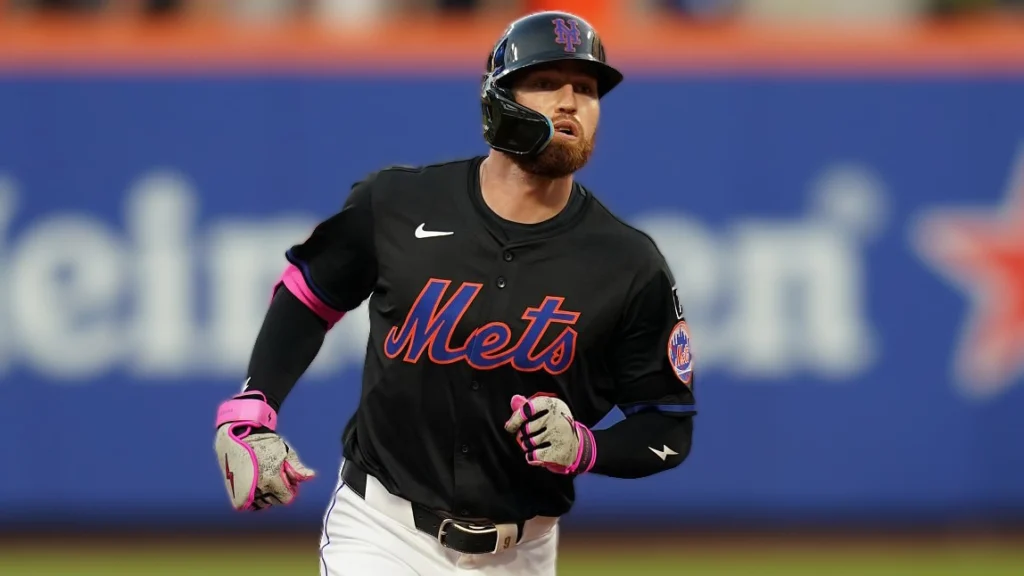 Brandon Nimmo, a baseball player for the New York Mets, wearing a black and pink Mets uniform, runs across the baseball field with a focused expression. He has a beard and is wearing a batting helmet. A blurred blue background with the "Heineken" logo is visible behind him.