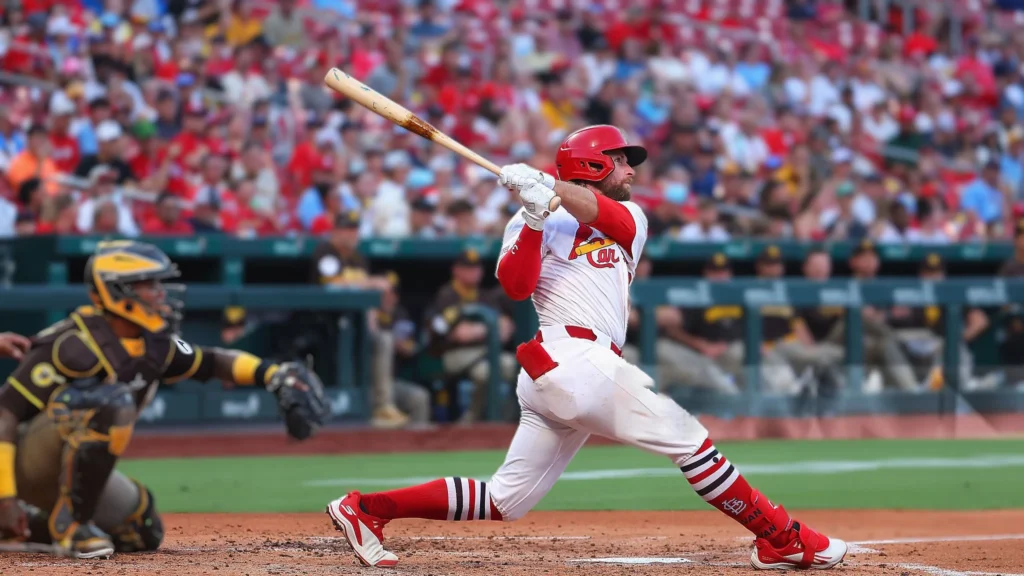 A baseball player in a white and red St. Louis Cardinals uniform, identified as Brendan Donovan, is captured mid-swing, having just hit a baseball. He is facing away from the viewer, looking towards where the ball would go. A San Diego Padres catcher in a dark and gold uniform is crouched behind him. In the background, a stadium filled with spectators is visible.