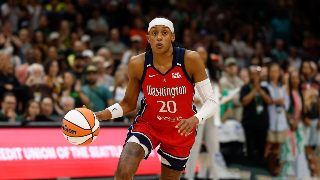 Brittney Sykes, wearing a red Washington Mystics jersey with number 20, dribbles a basketball while looking forward with determination on a basketball court. Spectators are blurred in the background.