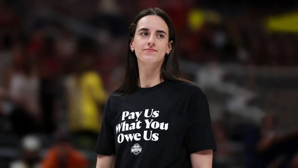 Caitlin Clark, a WNBA player, standing in an arena, wearing a black t-shirt with the WNBPA logo and the phrase "Pay Us What You Owe Us" in white text, representing player demands for fair compensation.