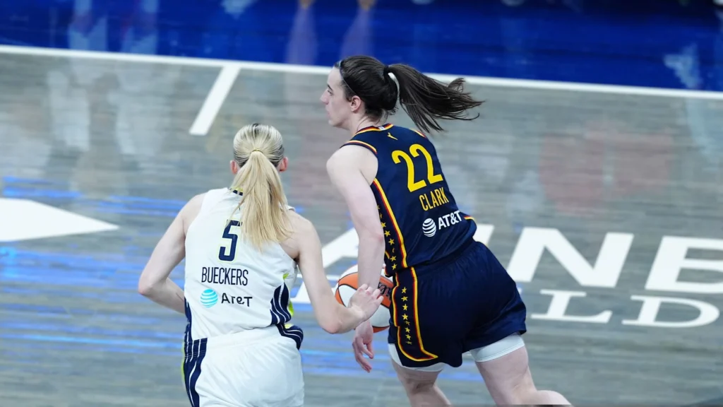 Caitlin Clark (Indiana Fever, #22) with the basketball, being guarded by Paige Bueckers (Dallas Wings, #5) on a basketball court.