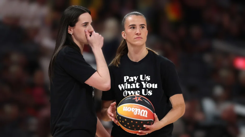 Sabrina Ionescu, a WNBA player, holds a basketball and wears a black t-shirt that clearly reads "Pay Us What You Owe Us" during a public appearance. Another player stands beside her.