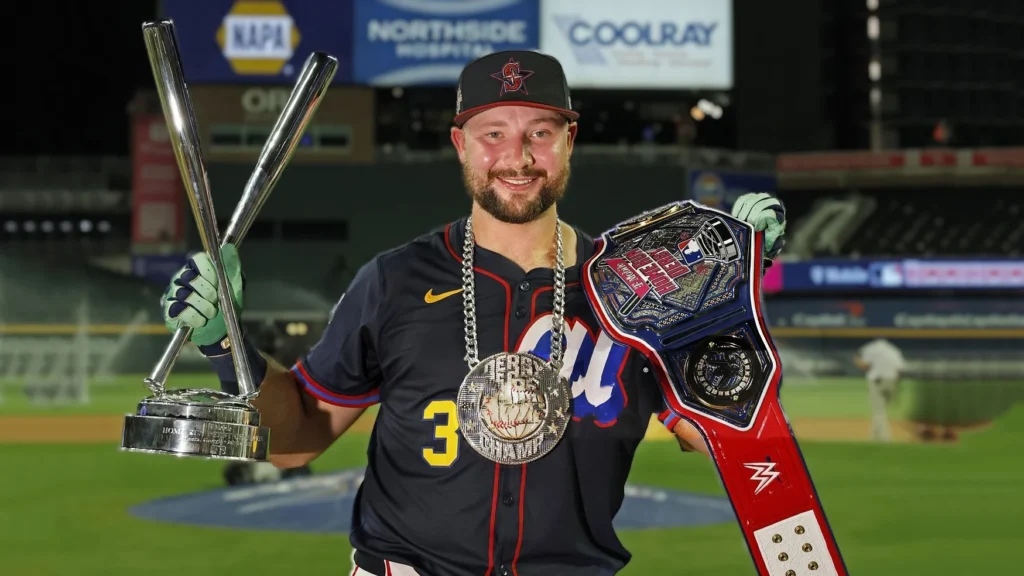 A smiling Cal Raleigh, a baseball player in a dark blue and red jersey and cap, stands on a baseball field at night, holding a silver trophy in his left hand and the large, championship-style Home Run Derby belt over his right shoulder. He is also wearing a thick silver chain with a large medallion around his neck. Stadium lights and advertising signs are visible in the blurred background.
