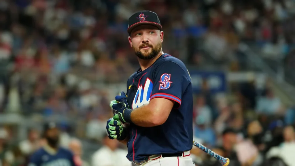 A close-up shot of baseball player Cal Raleigh wearing a navy blue jersey with red and white trim, a matching baseball cap, and green batting gloves, holding a bat and looking out onto the field. He has a beard and appears focused.