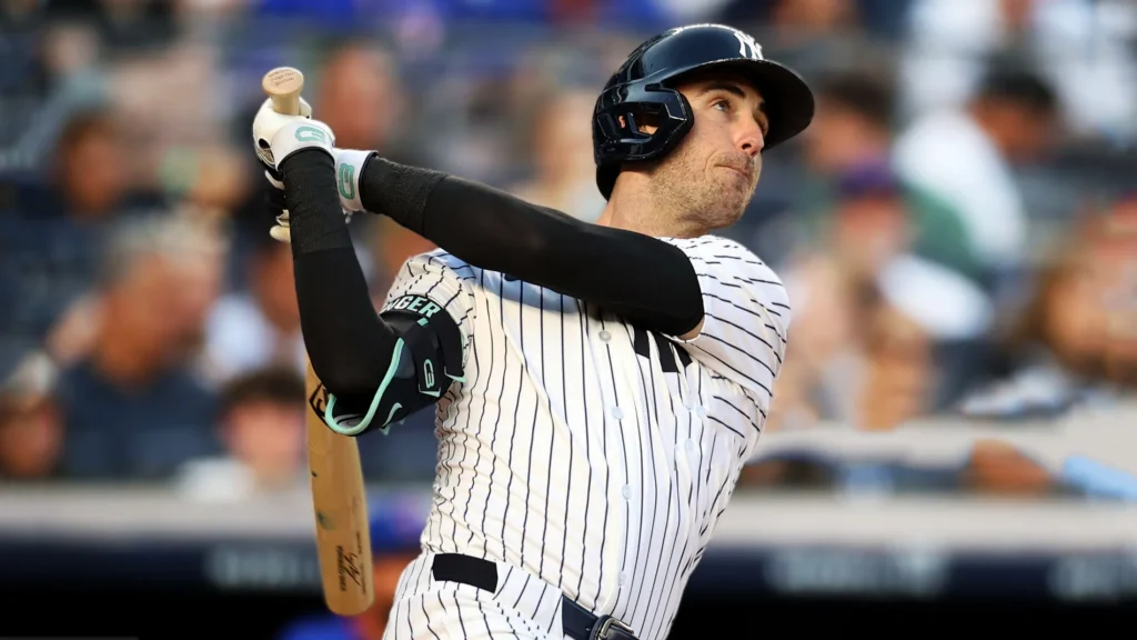 A close-up shot of New York Yankees player Cody Bellinger in a white pinstriped uniform and black helmet, mid-swing with a wooden baseball bat. He is looking intently towards the right, with a blurred stadium crowd in the background.