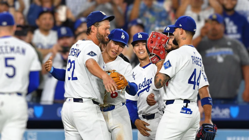 Clayton Kershaw, wearing a Dodgers uniform with number 22, is smiling and being embraced by his teammates on the baseball field. Shohei Ohtani (number 17) and Andy Pages (number 44) are visible among the congratulating players. The background shows blurred fans in a stadium, celebrating the moment.