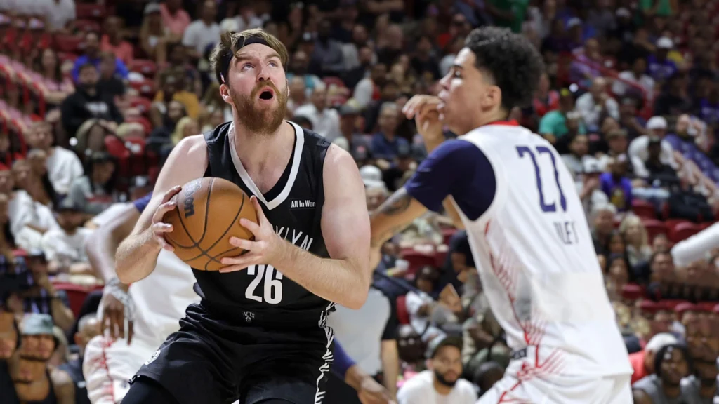 Drew Timme, a bearded basketball player in a black Brooklyn Nets jersey with the number 26, holding a basketball and looking up during a game. A Washington Wizards player in a white and blue jersey with the number 27 is defending him. The background shows a crowded basketball arena.