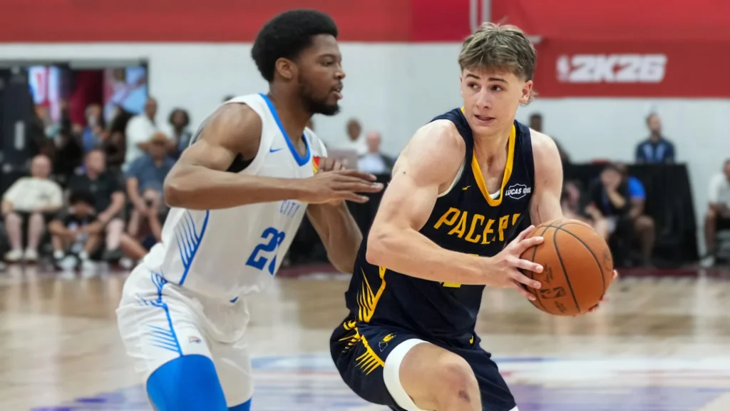  A basketball player, Johnny Furphy, wearing a blue and yellow Indiana Pacers jersey with "PACERS" written across the chest, dribbles the ball with his right hand while looking towards the basket. Another player in a white and light blue jersey with the number "21" defends him closely. The game is taking place on a basketball court with spectators in the background.