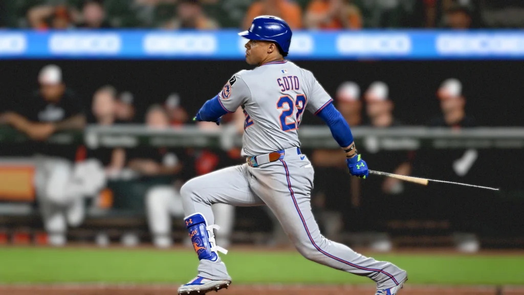 New York Mets outfielder Juan Soto in his batting stance, wearing a grey road uniform with "SOTO" and number "22" on the back, swinging a bat during a baseball game. He is looking towards the left of the frame, with a stadium background.