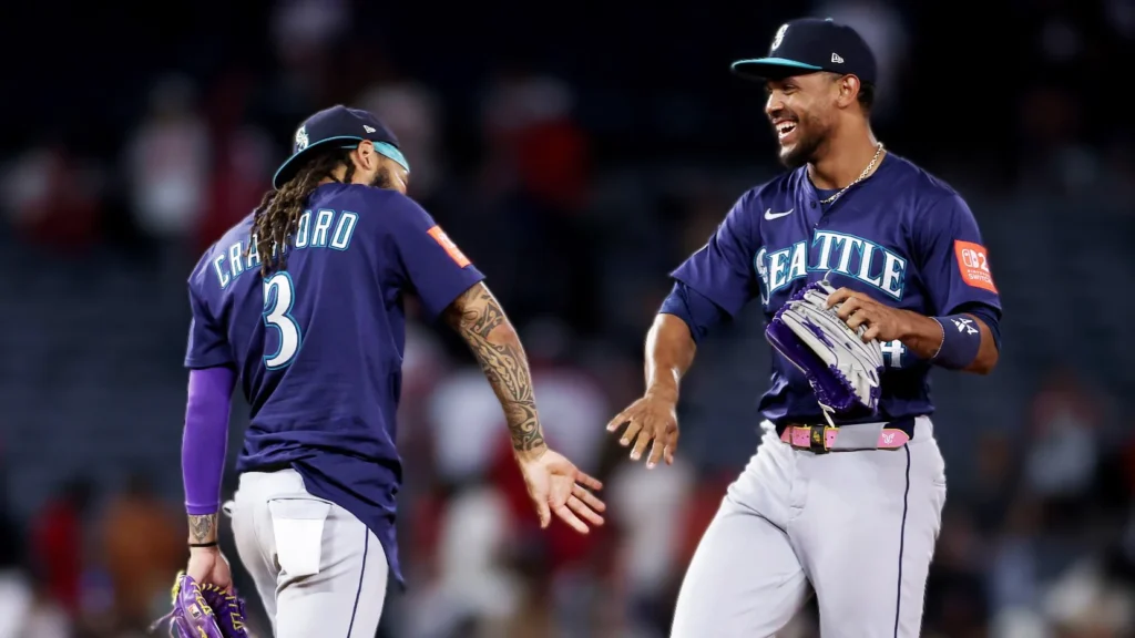Two Seattle Mariners players, J.P. Crawford (number 3, seen from behind) and Julio Rodríguez (smiling, facing forward, holding a glove), celebrating on the baseball field during a night game against the Los Angeles Angels. Both are wearing purple and grey Mariners uniforms.