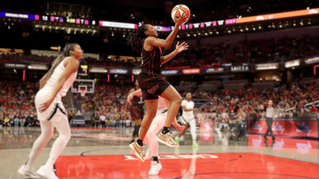 Kelsey Mitchell (Indiana Fever, #0) in mid-air taking a jump shot during a WNBA game. She is wearing a black and red uniform with gold shoes. A Las Vegas Aces player in a white uniform is nearby. The basketball court is red, and stadium lights and spectators are in the background.