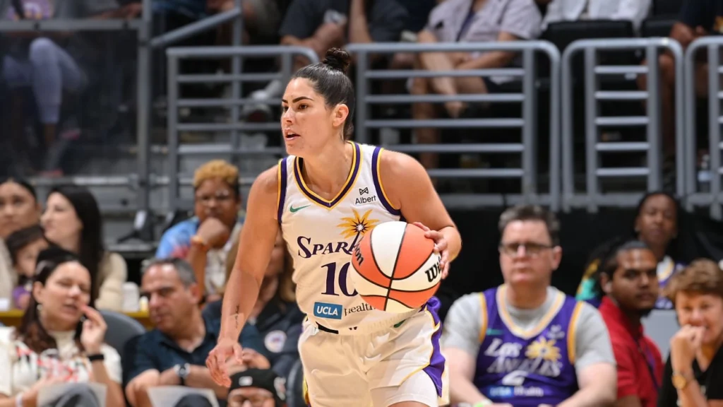 A close-up shot of Kelsey Plum, a female basketball player for the Los Angeles Sparks, in action on the court. She is wearing a white and purple Sparks uniform with the number 10, holding a basketball, and looking intently forward with a focused expression. Spectators are blurred in the background.