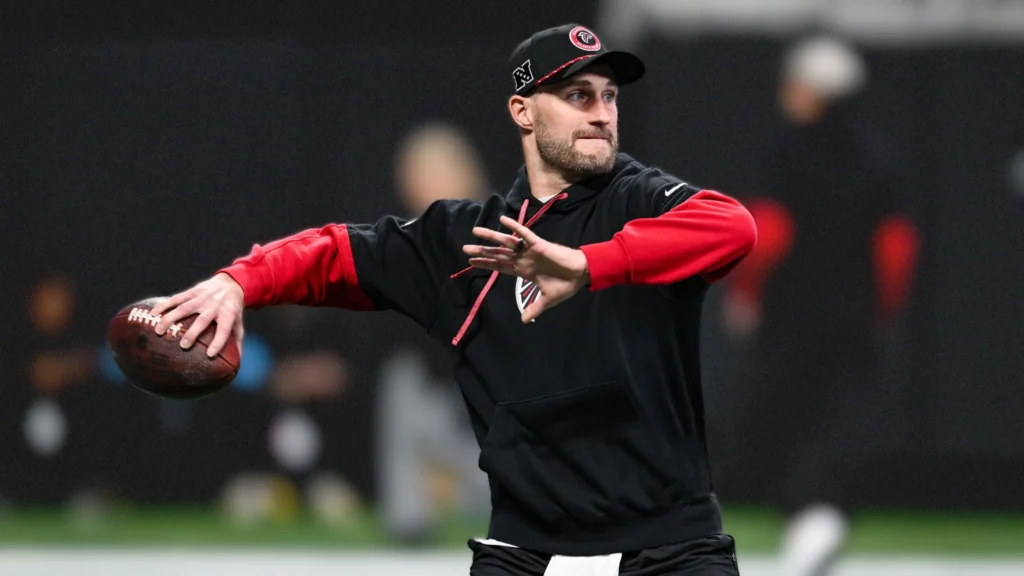Kirk Cousins, a white male quarterback with a beard, wearing a black and red Atlanta Falcons hoodie and a black baseball cap, is in the middle of a throwing motion with a football in his right hand. He is on a dark field.