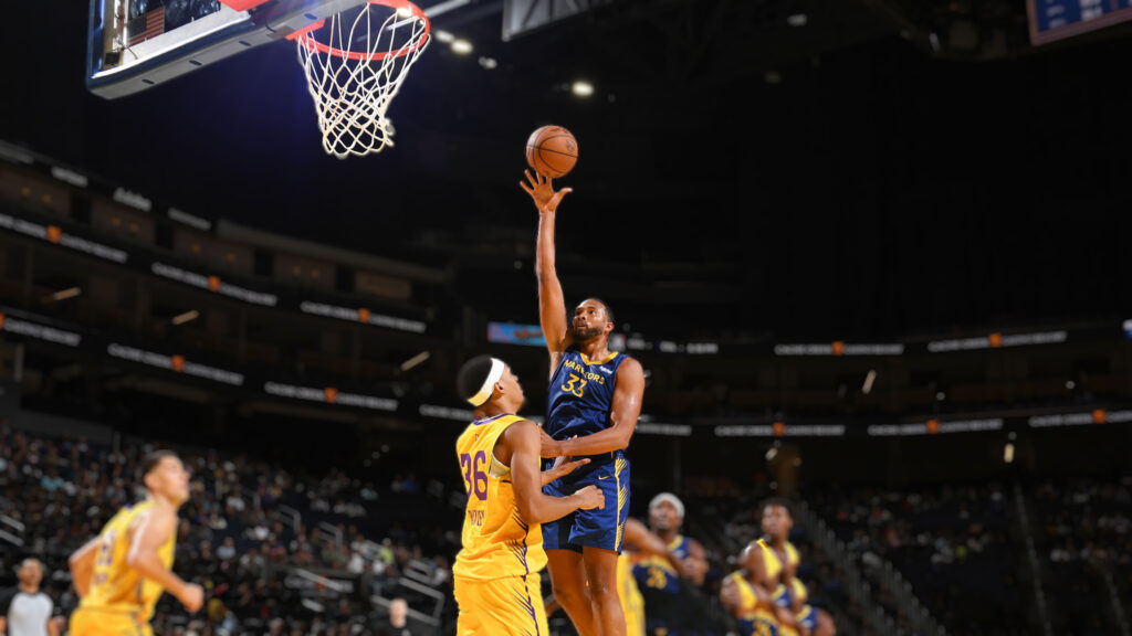Golden State Warriors player, wearing a blue and gold jersey with number 33, shoots a layup over a Los Angeles Lakers player in a yellow jersey with number 26, during an NBA Summer League game. The basketball hoop and a dark arena crowd are visible in the background.