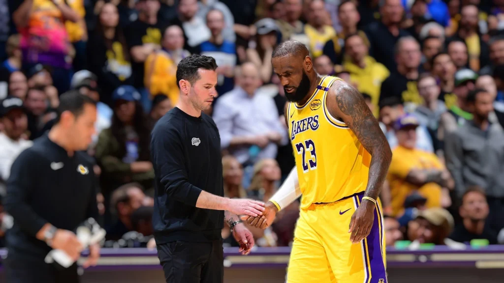 LeBron James, in a yellow Lakers jersey with "LAKERS 23" on it, shaking hands with a male team staff member in black attire on the basketball court sidelines. A blurred crowd is visible in the background.