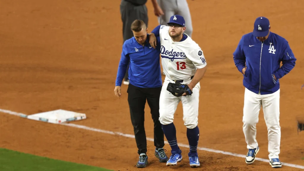 Max Muncy, a Dodgers player wearing jersey number 13, is being supported by two trainers as he limps off a baseball field, clearly in pain. He is wearing a white Dodgers uniform with blue accents. The ground is dirt, and a white base is visible behind him.