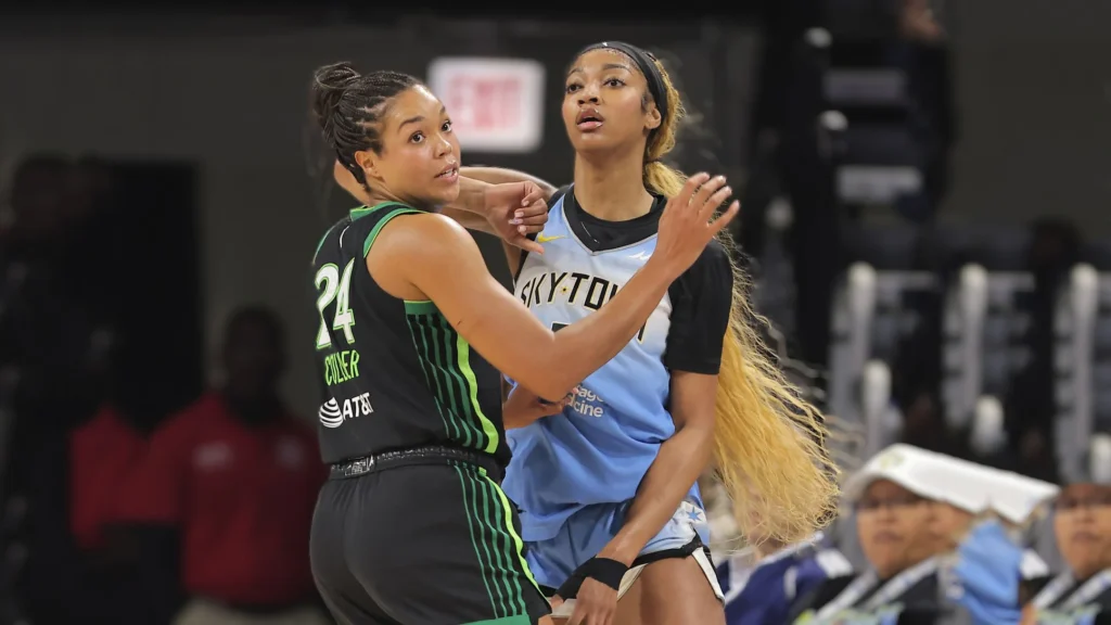A close-up action shot of Lynx vs Sky matchup, where two WNBA players: Napheesa Collier in a black and green Lynx jersey with the number 24 on her back, facing and defending against Angel Reese in a light blue Sky jersey. Both players are engaged in a physical struggle for the ball or position on the court.