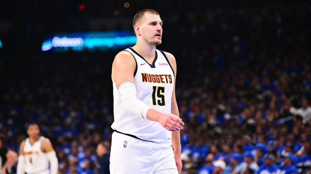 A basketball player wearing a white Denver Nuggets jersey with the number 15, looking to his right on a basketball court filled with spectators.
