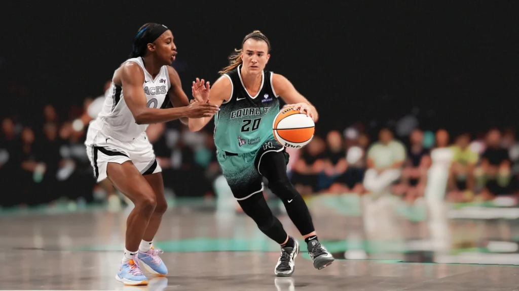 Sabrina Ionescu, wearing a seafoam green and black Liberty jersey with "EQUITY" written on it, dribbles a basketball with her right hand while being guarded by an Aces player in a white and black uniform. Both players are in motion on a basketball court.