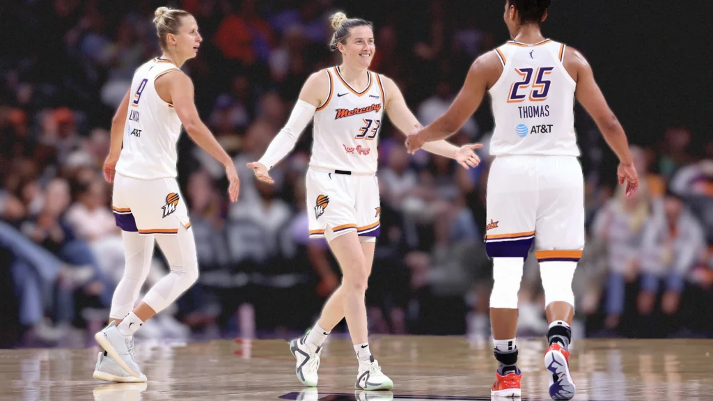 Three Phoenix Mercury basketball players in white uniforms celebrating on the court. The player in the center, Sami Whitcomb (#33), is smiling and has her arms slightly outstretched. Another player (#9) is on her left, and Alyssa Thomas (#25) is on her right.