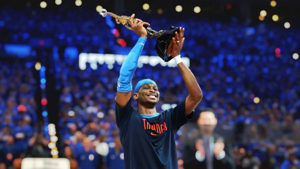 Shai Gilgeous-Alexander, wearing an Oklahoma City Thunder jersey and blue headband, smiling broadly and holding up the golden NBA championship trophy with both hands. He is on a basketball court with a blurry blue crowd in the background.