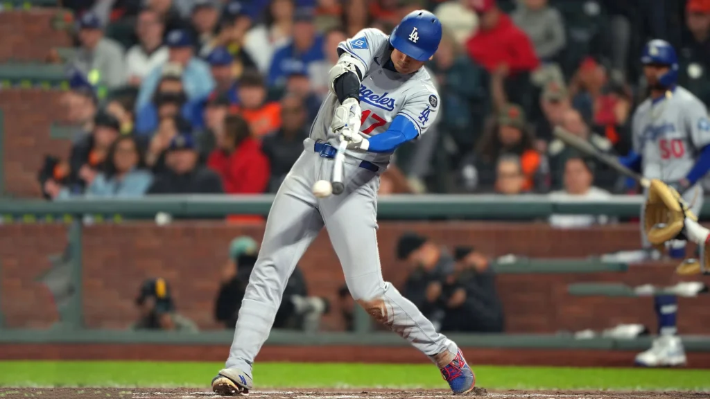 Shohei Ohtani, wearing a gray Los Angeles Dodgers uniform with a blue batting helmet, swings hard at a baseball, making contact in mid-air. He is in the batter's box at Oracle Park with a blurred background of fans and the opposing San Francisco Giants player (catcher or umpire) in the background.