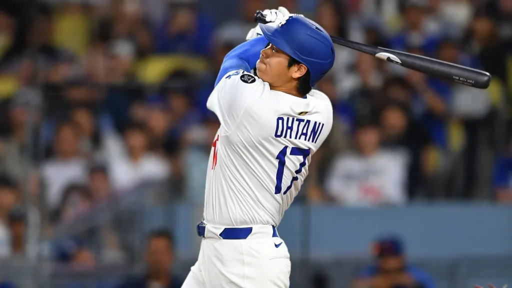 A close-up, action shot of Shohei Ohtani, wearing a white Dodgers uniform with "OHTANI" and number "17" on the back, in the middle of a powerful baseball swing at Dodger Stadium. His eyes are focused on the ball as the bat follows through, with blurred spectators in the background.