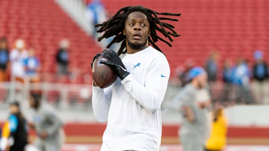 A close-up shot of NFL quarterback Teddy Bridgewater, with dreadlocks, wearing a white long-sleeved Detroit Lions shirt and black gloves, holding a football on a red stadium field. He looks focused with a serious expression.