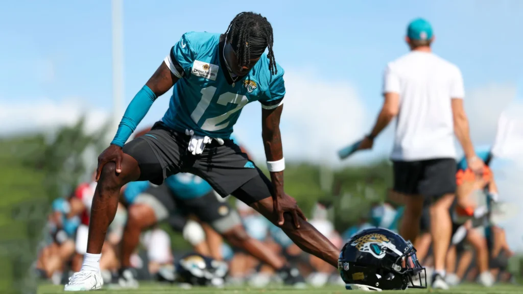 Travis Hunter, a Black male athlete with dreadlocks, wearing a teal Jacksonville Jaguars jersey with the number 12 and black shorts, stretches his right leg out to the side with his torso bent forward. He is on a sunny green field, and his black Jaguars helmet rests on the grass near his right foot. Other blurred figures and a coach are in the background.