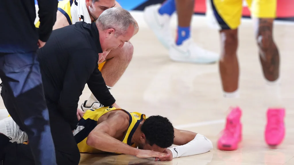 Tyrese Haliburton, wearing a yellow Pacers jersey, lying face down on the basketball court in pain with medical staff kneeling beside him.