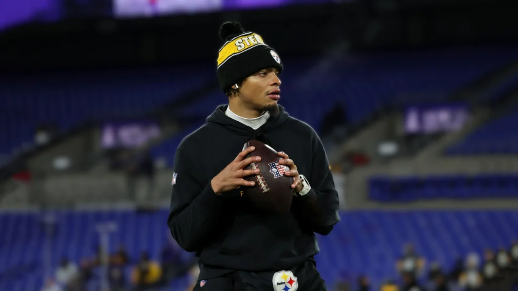 Justin Fields, wearing a black hoodie and a black and yellow knit hat with "STEELERS" written on it, holds a football on a blurred purple stadium background.