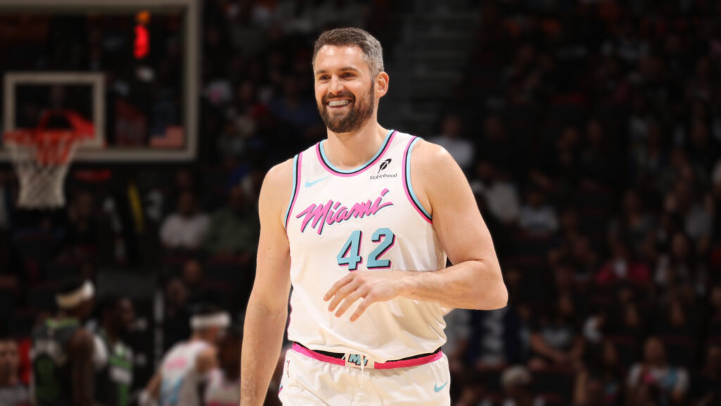 smiling Kevin Love, a white male basketball player with a beard, wearing a white Miami Heat jersey with "Miami" and the number 42 in pink and light blue, stands on a basketball court with a blurry audience in the background.