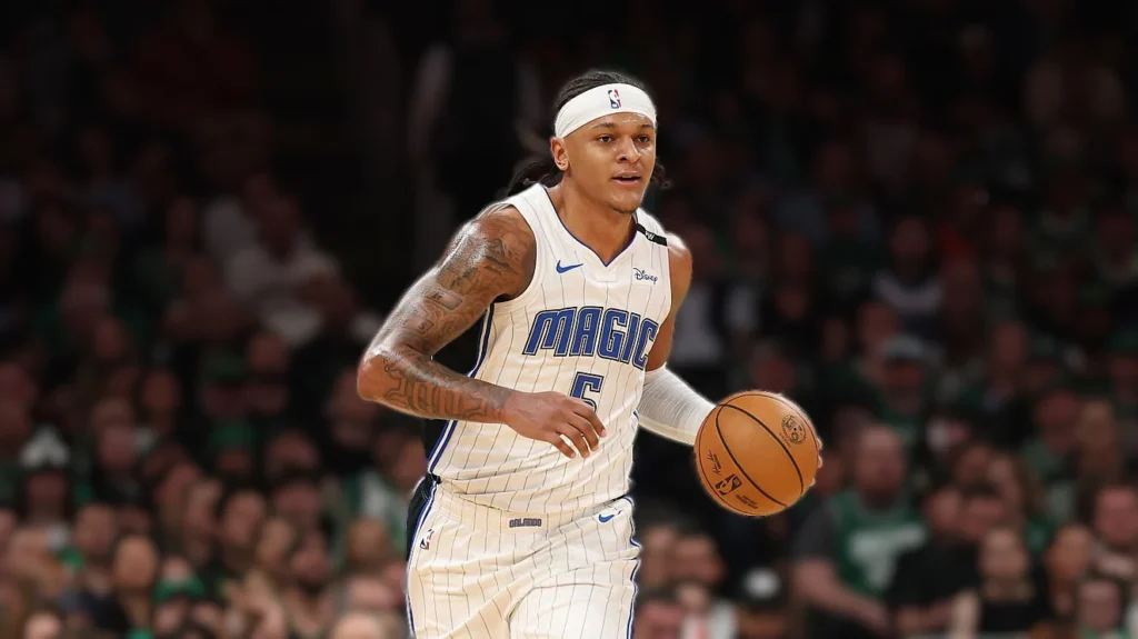 Paolo Banchero, wearing an Orlando Magic white jersey with blue trim and a white headband, dribbles a basketball with his right hand, looking towards his left. He has braided hair and visible tattoos on his arms. The background is a blurred view of a basketball arena crowd.